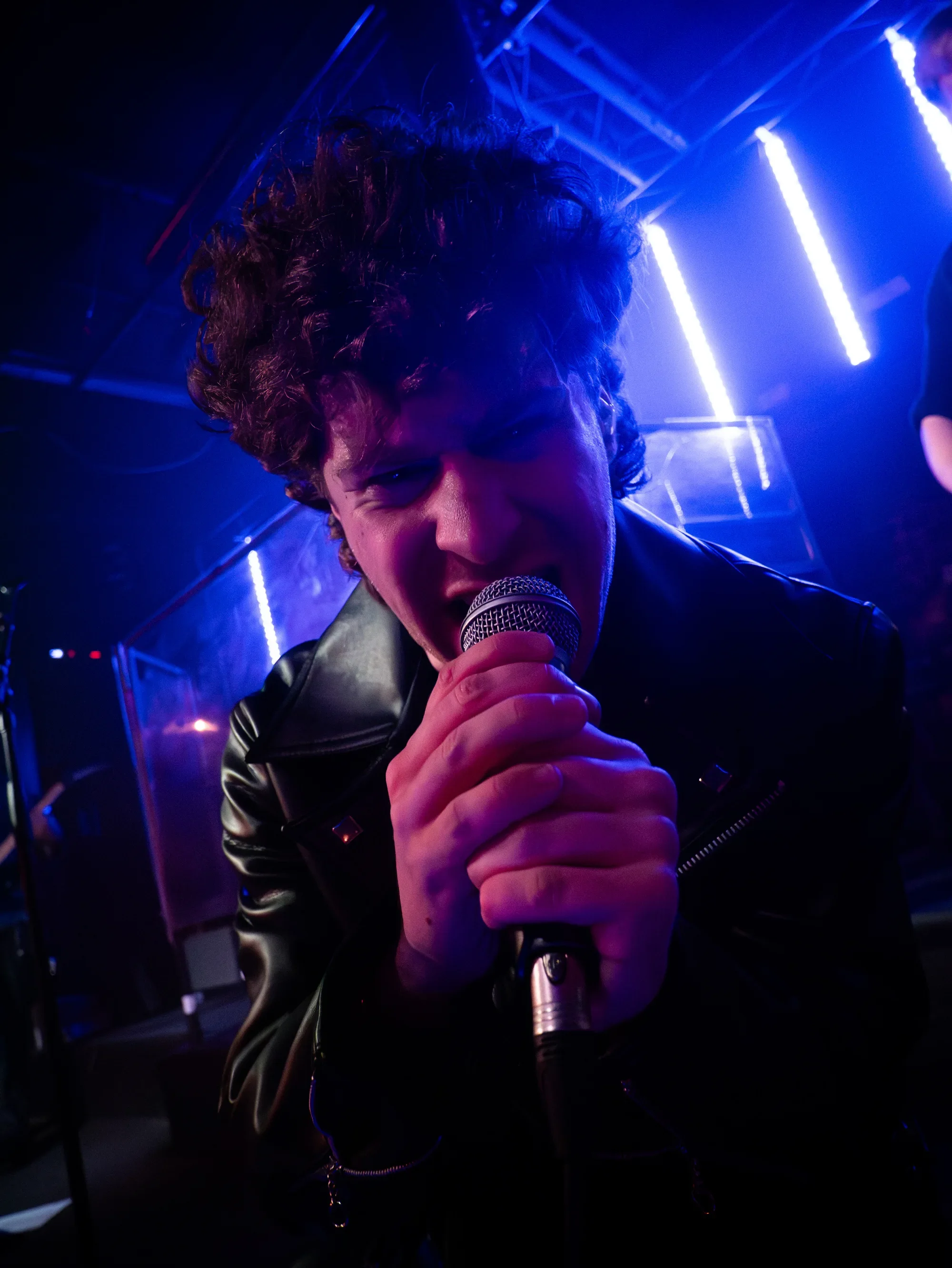 MAGNETIQ vocalist leaning towards the camera while singing into a microphone under blue and purple lights.